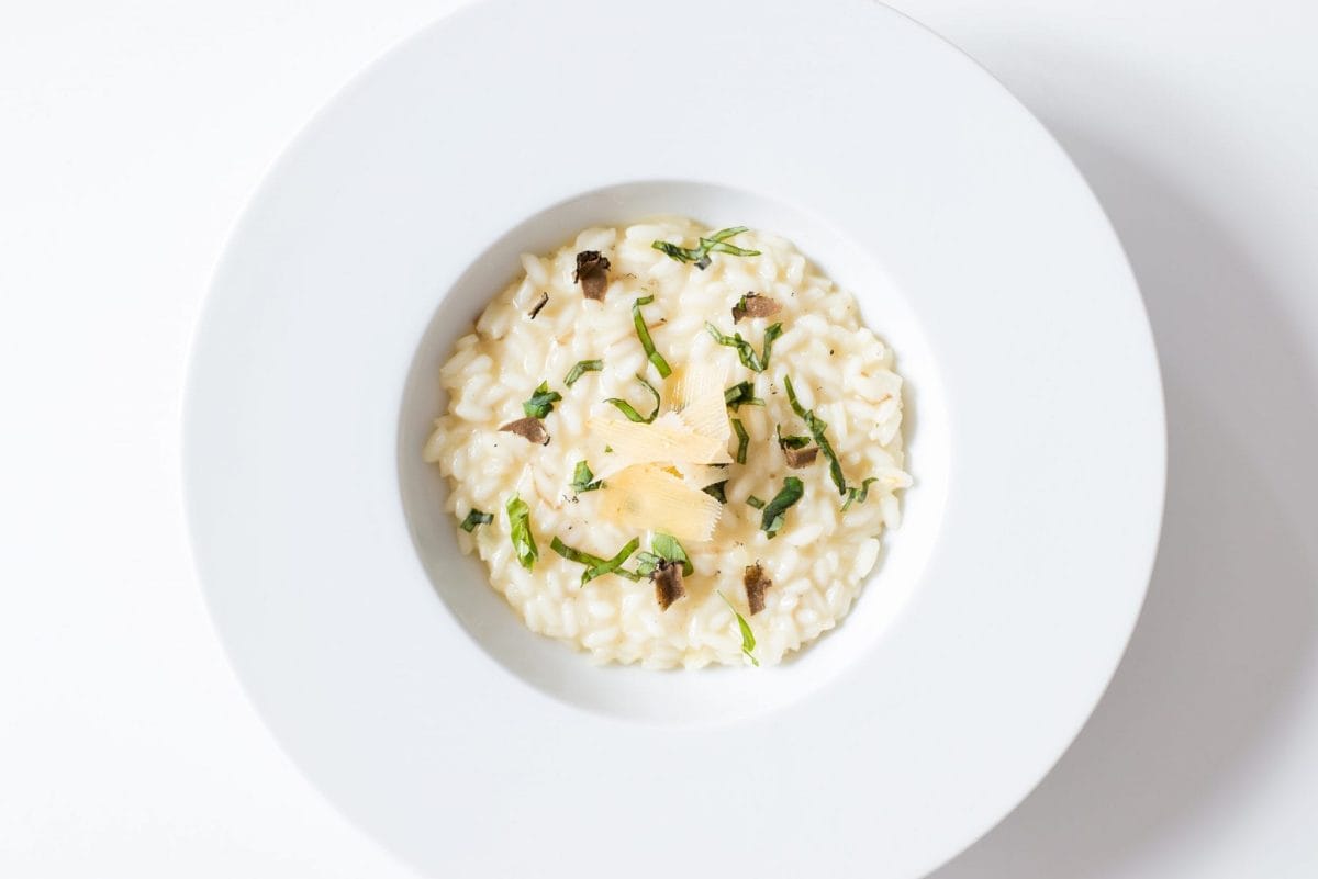 Plate of white risotto in a white bowl topped with shavings of cheese, truffle, and herbs