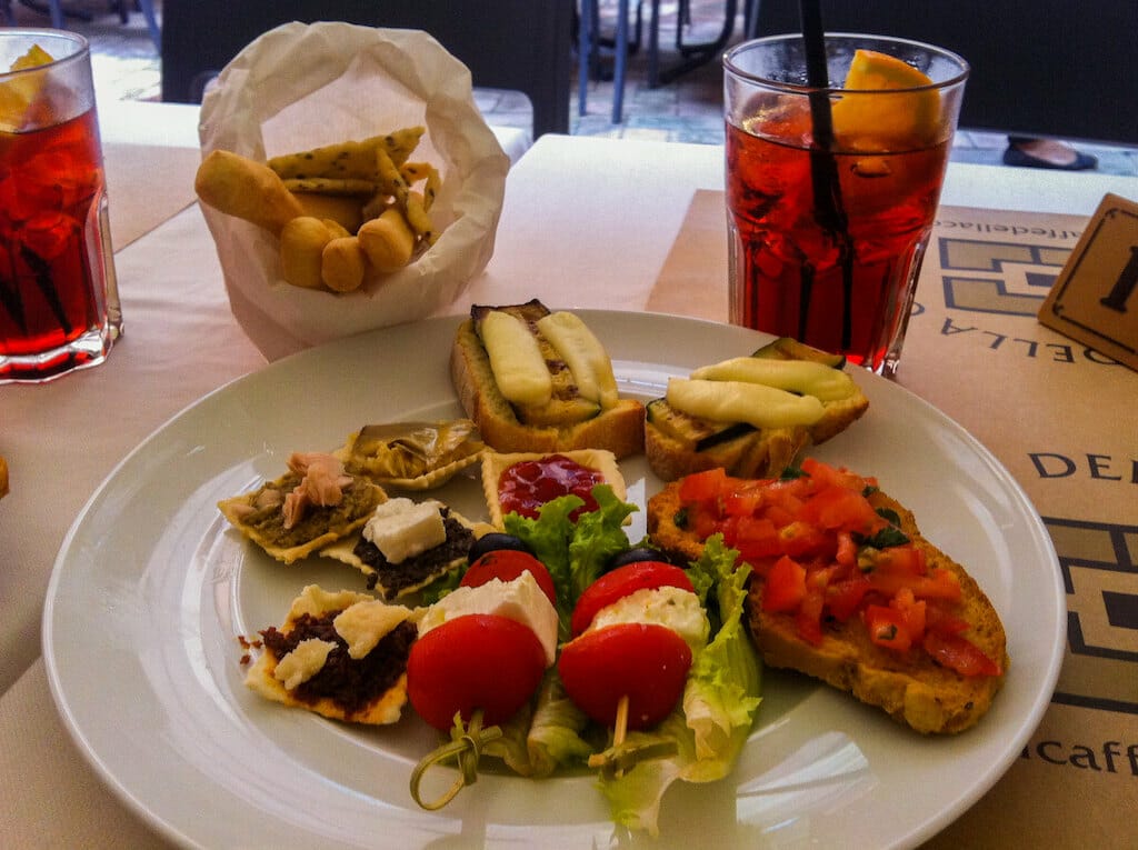 Aperitivo spread from Bologna, Italy, which includes a Campari, bruschetta, grisini, zucchini toast, cherry tomatoe bites, and mozzarella.