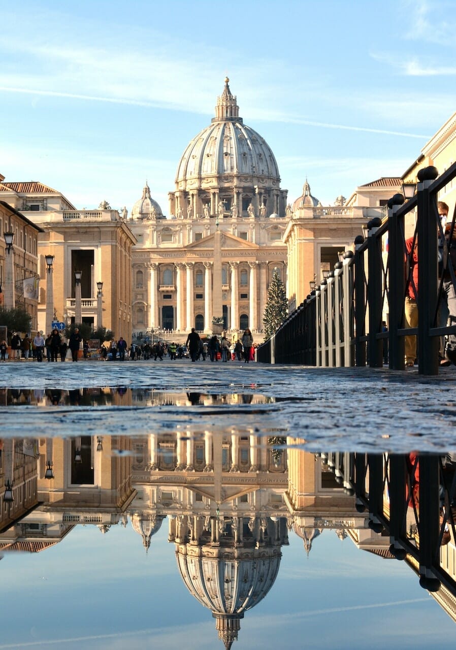 Vertical photo of the Vatican and St. Peter's Basilica with water puddle reflecting the dome and lines of crowds